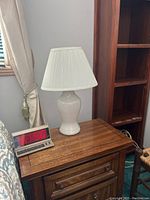 Pair of white ceramic table lamps shown on wooden furniture, each with off-white pleated fabric lampshades.
