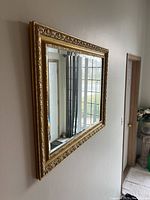 Side angle view showing the thickness and detailed carvings of the gold frame and the mirror reflecting a window and door.