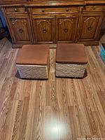 Pair of woven foot stools with brown cushioned tops placed side by side on wood floor in front of a wooden cabinet.