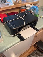 Black Epson all-in-one inkjet printer on kitchen counter with power cord draped over it and paper loaded in tray.