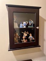 Front view of dark brown wooden display shelf with glass door showing various small ceramic and porcelain figurines and pitchers displayed inside.