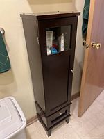 Small dark wooden cabinet with glass panel door and drawer, shown fully closed with items visible through glass.