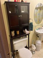 Front view of dark wooden toilet caddy shelf mounted over toilet tank with upper cabinet doors and lower open shelf holding three items.