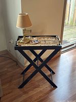 Photo showing black wooden folding TV tray with removable decorative tray, brass table lamp, and small round plate on tray.