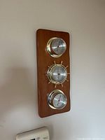 Full view of vertical wooden plaque with three brass-weather instruments mounted: thermometer top, barometer middle, hygrometer bottom.