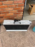 Folded aluminum wheelchair ramp resting against a brick wall outdoors on a concrete surface. The ramp has a black non-slip surface and a carrying strap.