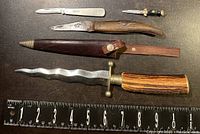 Four vintage cutting tools arranged in a row with measuring ruler, showing wavy blade knife, folding knives, and mini spring cutter.