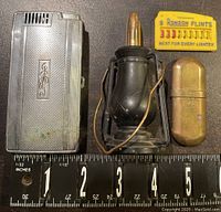 Three vintage lighters including a rectangular silver-tone lighter with initials, brass and black lantern style lighter, and brass capsule lighter with 'BRASS' marking alongside a Ronson flints pack and ruler.