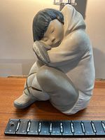 Front and side views of Lladro porcelain figurine of indigenous child sitting with eyes closed, detailed hood, mittens, and boots with brown accent lines. Includes ruler for scale.