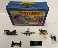 Matchbox carry case with six diecast vehicles laid out in front, includes Y13 American locomotive, Y8 Sunbeam motorcycle, Dinky seaplane, Hot Wheels Batmobile, open wheel racer, and vintage car.
