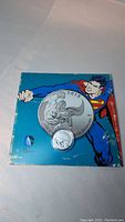 Photo of the coin in protective case on top of blue card backing with Superman image and DC Comics logo.