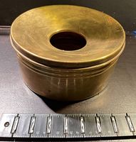 Side angle showing round brass spitoon with ruler indicating about 4 inches height and circular shape.