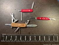 Three assorted multi-use Swiss made pocket knives arranged on table with ruler for scale.