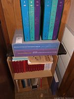 Overview of wooden shelf showing colored binder sets on top shelf, slipcases and loose pamphlets on middle shelf, red Torch Bearer volumes on bottom shelf