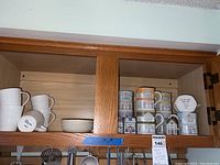 Cabinet shelf showing plain white mugs, small bowl, Lenox Village mugs and house-shaped spice jars