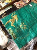 Stack of green and black T-shirts in plastic bag showing green shirt on top with gold Nagano 1998 torch relay graphic
