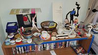 Shelf overview showing assorted Olympic-themed tins, trinket boxes, porcelain figurines, decorative bowl, pewter plate, model bus, cup and saucer set