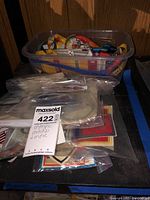 Clear plastic bin filled with multicolor woven lanyards and sealed bags of embroidered patches on black shelf