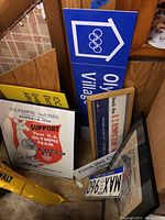 Photo showing framed 1956 Australia Olympic Games poster, blue Olympic Village sign with rings, a metal license plate, and other signs leaning against wood panel wall on parquet floor.