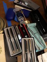 Photo showing multiple pen sets including three boxed Quill pen sets, additional pens in Tiffany & Co pouch and other cases, plus a wristwatch and NBC watch pendant