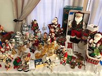 Wide view of many Christmas themed holiday figurines and ornaments laid out on a white surface including multiple Santa Claus, snowmen, elves, reindeer, and other festive figures. Includes boxed items and loose figures with some gold accents and colorful clothing.