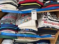 Photo of folded t-shirts with visible Olympic rings and various sports prints, showing multiple stacked items on shelf