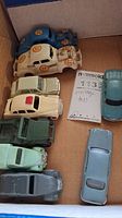 Top-down view of 9 vintage toy cars in a cardboard box showing a variety of colors and conditions, including blue, cream and green models. Some cars have numbered decals.