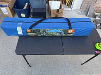 Packaged Ozark Trail canopy in a blue bag with black straps, laid on a black folding table outdoors.