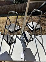 Two metal step stools standing unfolded on table outdoors with shadows cast.
