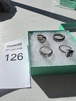 Four silver rings displayed in green box with white padding; two rings with purple gemstones, one with clear stone, and one ornate band.