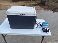Closed view of the Coleman electric cooler on white table showing the blue body, white lid, and associated cords and transformer.