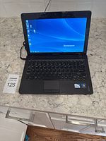 Lenovo Ideapad laptop powered on showing Windows desktop on screen, placed on kitchen countertop with charger connected.