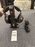 Front view of Sennheiser HDR 185 wireless headphones on charging stand with power adapter cable visible to the right.