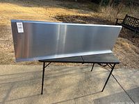 Full frontal view of stainless steel shelf resting on a black folding table outside, showing length and backsplash.