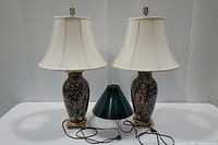 Full frontal photo showing pair of porcelain lamps with white shades and separate dark green shade in between; dark green glaze with floral motifs and brass bases visible.