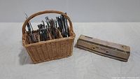 Woven utensil basket filled with assorted flatware and a boxed item beside it on a white cloth.