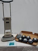 Front view of the full Electrolux floor scrubber/polisher unit with chrome handlebars and beige body, six round brush heads in a tray, and owner's manual laid out on a table.