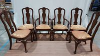 Six brown solid wood dining chairs arranged in two groups, showing deep carved details on the backrests and curved legs with pad feet. Upholstered seats are gold with red floral and dragon design.