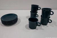 5 teal green-blue Fiesta Ware mugs standing and stacked in two columns beside 6 stacked matching saucers on white background