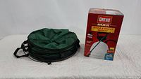 Green collapsible waste container collapsed with carrying straps visible next to boxed Ortho MAX Weed & Insect Sprayer.