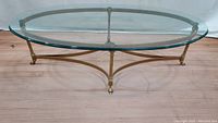 Full side view of the oval glass top coffee table showing the brass frame and claw feet on wooden floor.