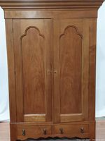 Front view of closed armoire showing light wood with arched panel doors and two hanging drawers below with ebony and brass drawer pulls.