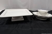 Photo showing the white square pedestal cake stand and the white double clam shell chip and dip bowl side by side on a black fabric background.