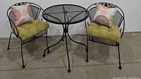 Photo showing two wrought iron cushioned patio chairs with decorative pillows and a round wrought iron mesh top patio table between them, all set on a concrete floor.