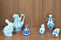 Six assorted blue and white ceramic items displayed on wooden background: rabbit figurine, floral pitcher, decorative bell, child figurine, dual-handled vase, shoe-shaped item.