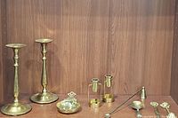 Entire lot displayed on wooden shelf: two tall brass candlestick holders, two small brass candle holders with handles, brass candle snuffer, brass letter opener, small round brass trinket box with hinged lid.