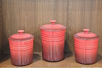Three cerise red Le Creuset stoneware canisters with lids displayed side by side on a wooden shelf