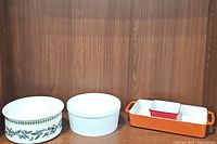 Four casserole dishes displayed on wooden shelf: Portmeirion Botanic Garden ceramic dish, white fluted ceramic dish, red ceramic portion dish nested in orange enameled cast iron tray.