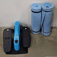 Blu compact seated elliptical trainer machine with black pedals and digital display, alongside two rolled blue exercise mats placed vertically.