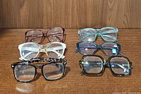 Six pairs of prescription or reading glasses arranged upright on a surface against a wood background showing various frame styles and colors.